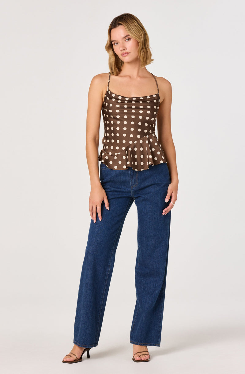A woman is wearing a brown sleeveless top with thin spaghetti straps and white polka dots. The top has a ruffled hem that flares out slightly at the waist. She is also wearing high-waisted, straight-leg blue jeans and beige open-toe high-heeled sandals.