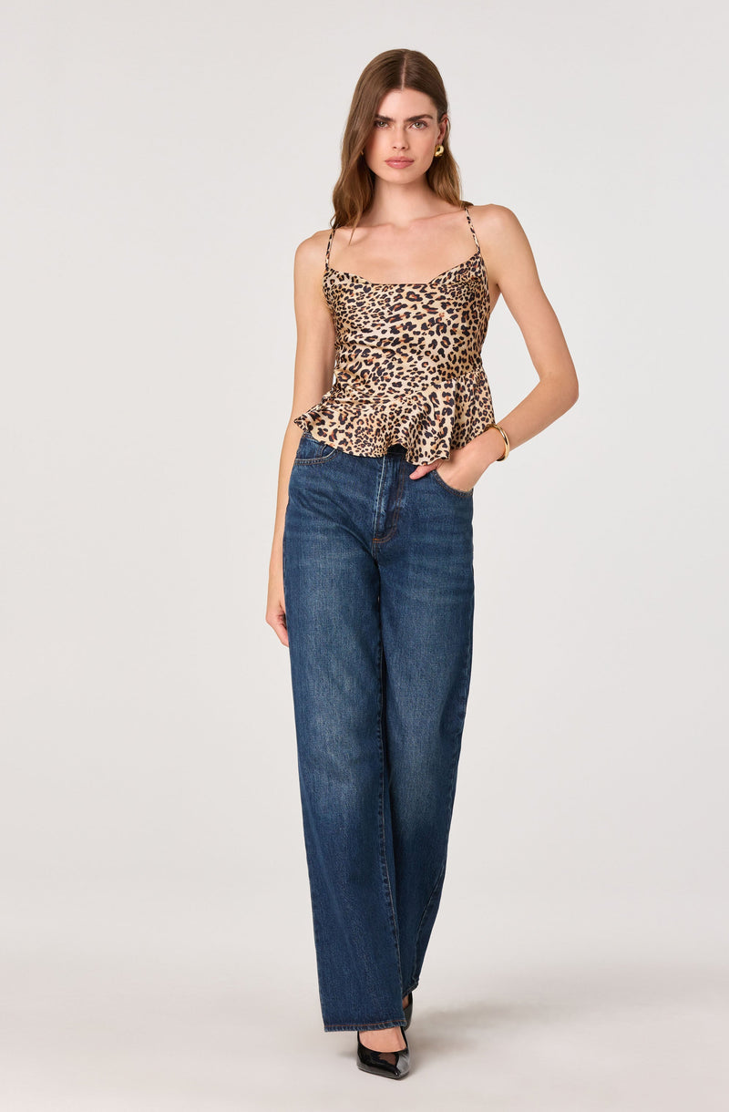 A woman is wearing a sleeveless, leopard print top with thin spaghetti straps and a ruffled hem. She is also wearing high-waisted, wide-leg blue jeans and black pointed-toe shoes. She has minimal jewelry, including small earrings and a bracelet.