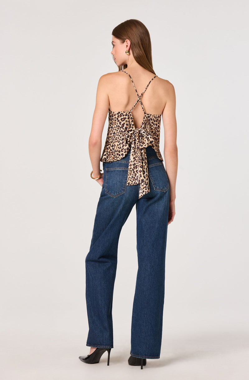 The woman is wearing a sleeveless, leopard print top with thin spaghetti straps that crisscross at the back and tie into a bow. She is also dressed in high-waisted blue jeans with large back pockets, paired with black high-heeled shoes.