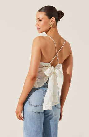 A woman is wearing a delicate, sleeveless, white lace top with thin spaghetti straps and a large bow detail at the back. She pairs it with light blue high-waisted jeans. She has dark hair styled in a sleek bun and is accessorized with large, gold earrings.