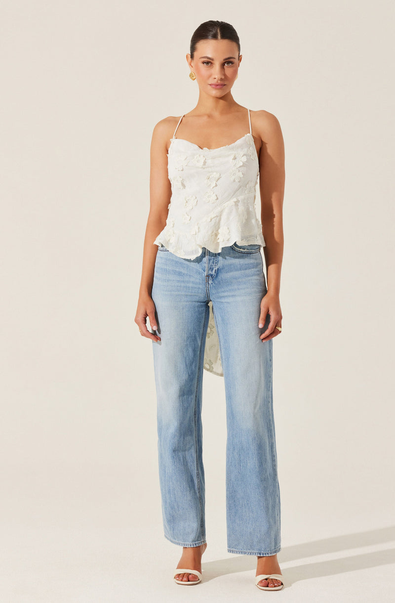A woman is wearing a cream-colored sleeveless top with spaghetti straps and a ruffled, textured front. She pairs this with light-wash, high-waisted, straight-leg jeans. She is wearing open-toed heels and has her dark hair styled in a sleek, pulled-back look.