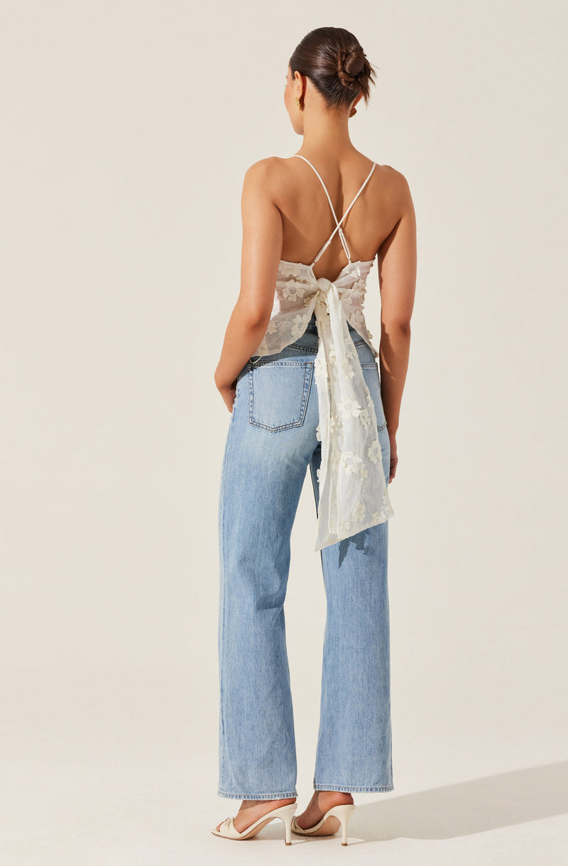 A woman is wearing a white lace top with thin spaghetti straps that crisscross at the back and tie into a large bow at the waist. She is also wearing high-waisted, light-wash denim jeans with a straight-leg cut. Her shoes are beige high heels with a pointed toe and ankle strap.