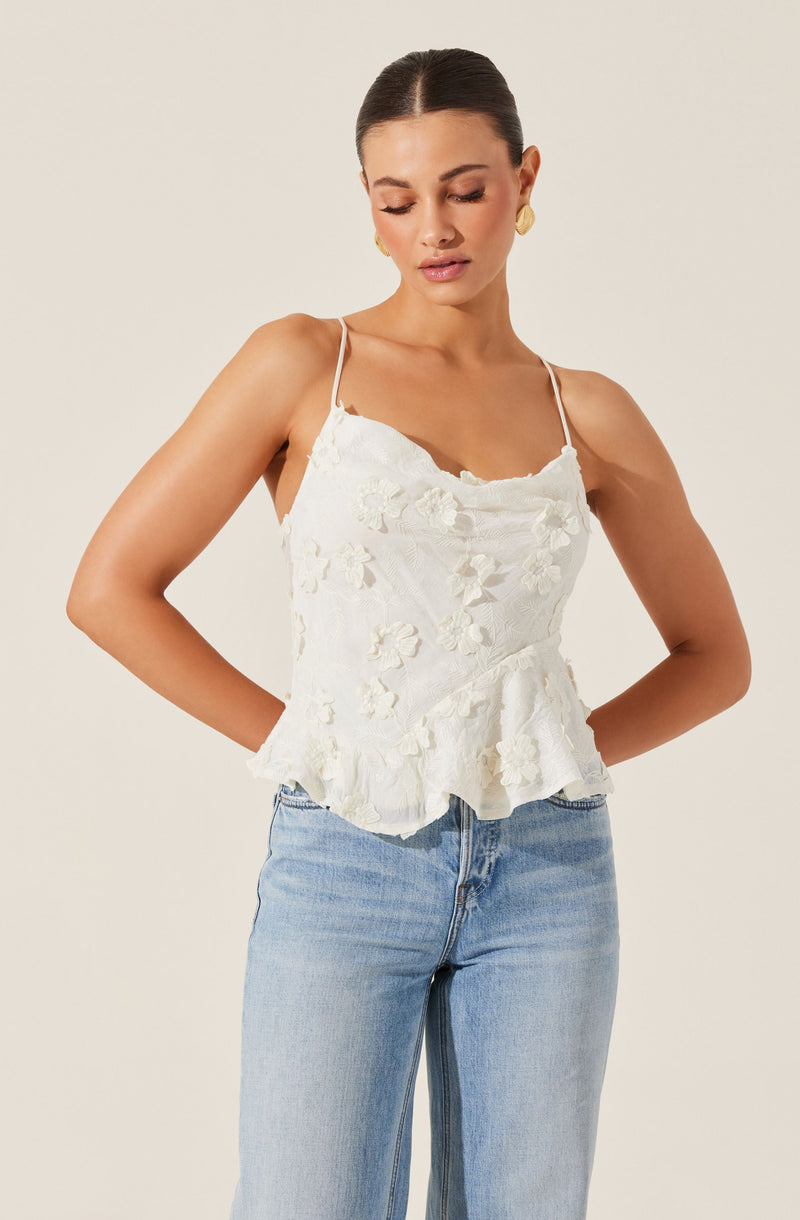 A woman is wearing a white sleeveless top with thin spaghetti straps, featuring floral embroidery and a ruffled hem. She pairs the top with light blue high-waisted jeans.