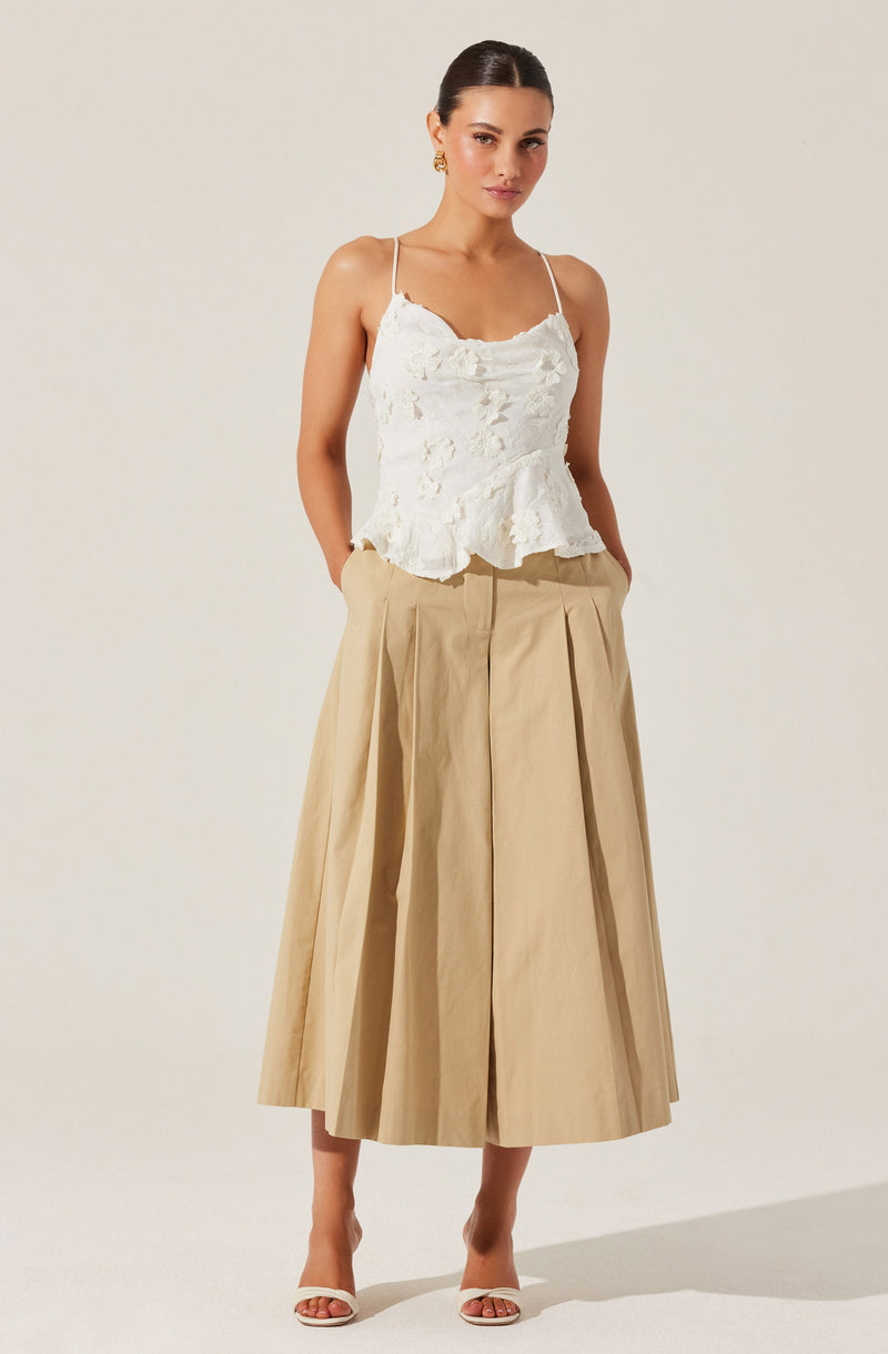 A woman is wearing a white sleeveless top with thin straps and a ruffled, textured fabric. She has paired it with a high-waisted, beige, wide-leg skirt that falls below her knees, with pleats adding volume. She is also wearing open-toed, beige high-heeled sandals.