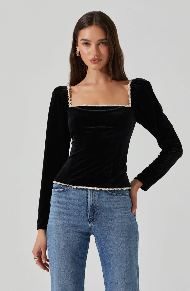 A young woman is wearing a black velvet long-sleeve top with a square neckline that is decorated with a white ruffled trim. The top fits snugly and has slight puffiness at the shoulders. She pairs it with light blue high-waisted jeans.