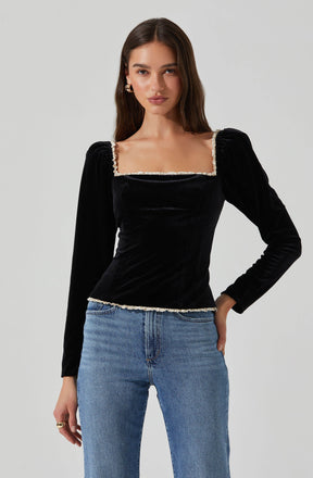 A young woman is wearing a black velvet long-sleeve top with a square neckline that is decorated with a white ruffled trim. The top fits snugly and has slight puffiness at the shoulders. She pairs it with light blue high-waisted jeans.