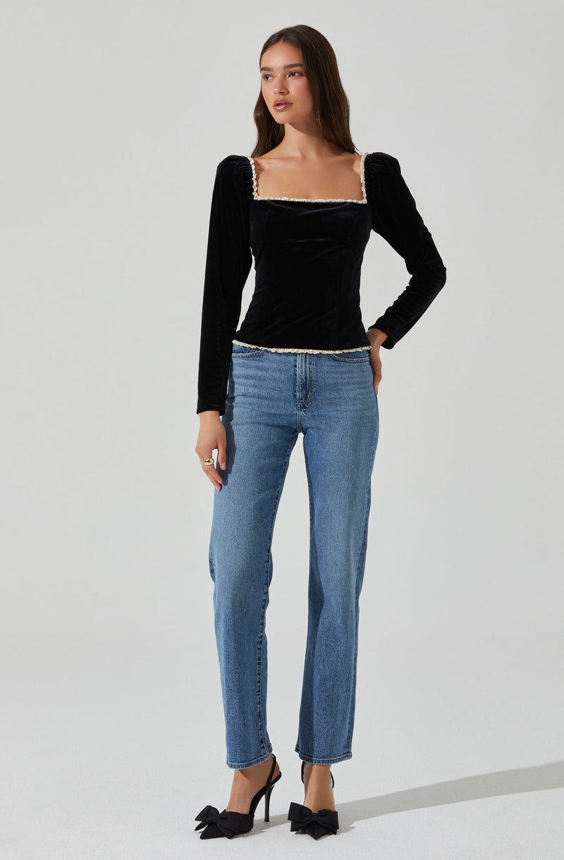 A woman is wearing a black, long-sleeved, velvet top with a square neckline accented by a decorative trim. She has paired the top with light blue, high-waisted, straight-leg jeans and black high-heeled shoes with bows on the front.
