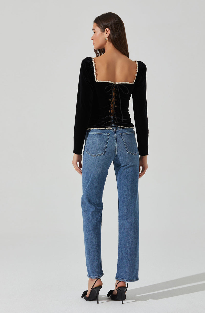 A woman is wearing a black long-sleeve top with a square neckline, decorated with a beaded trim, and a lace-up back detail. She pairs the top with high-waisted, straight-leg blue jeans and black high-heeled shoes with ankle straps.