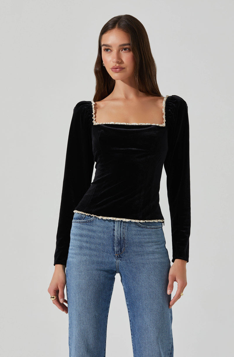 A woman wearing a black velvet long-sleeve top with a square neckline, decorated with a light-colored, possibly beaded, trim along the edges of the neckline and hem. She pairs the top with high-waisted blue jeans.