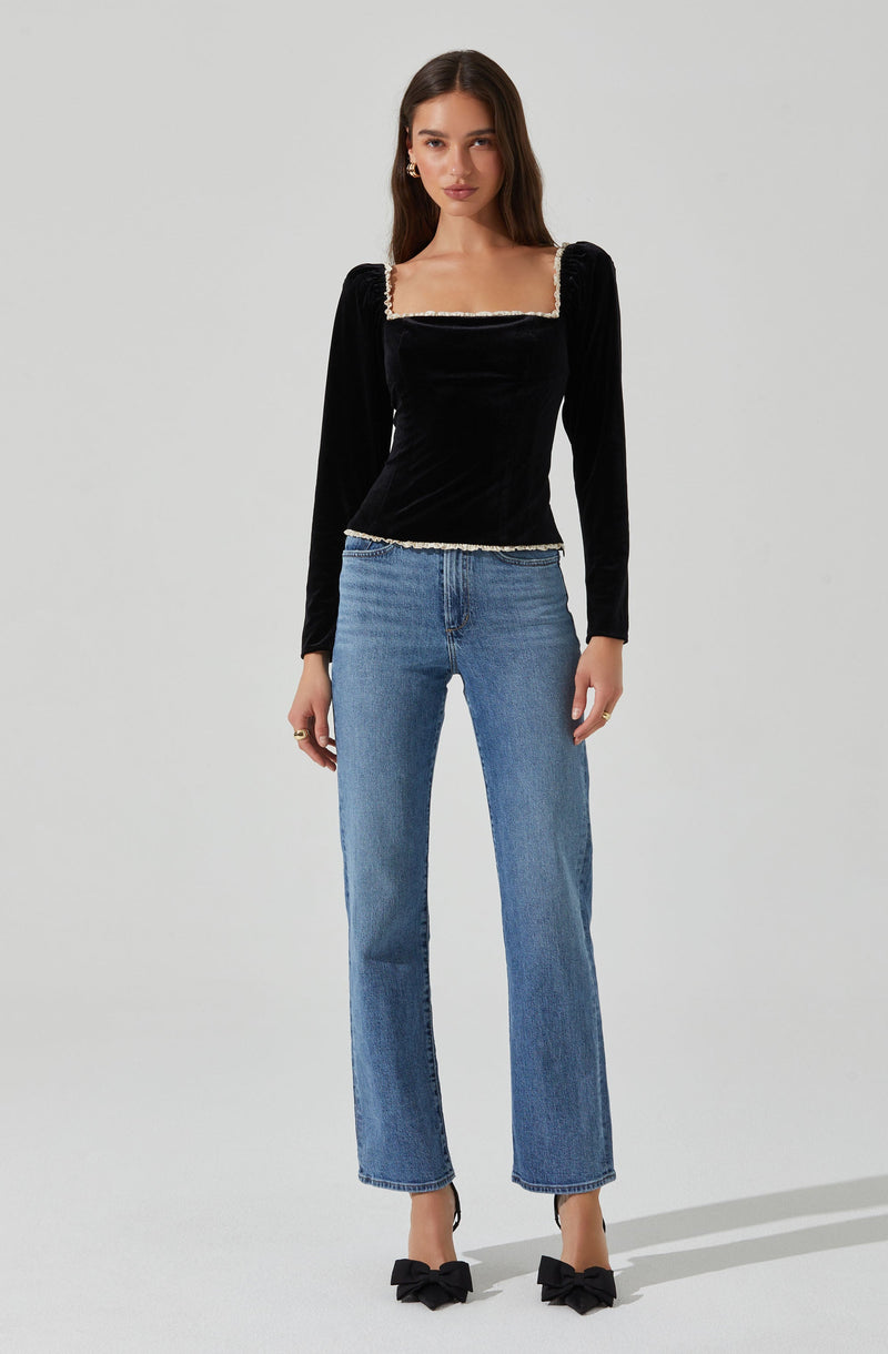 A woman is wearing a black velvet long-sleeve top with puffed shoulders and a square neckline, accented with a decorative trim along the neckline and hem. She pairs the top with light blue straight-leg jeans and black shoes adorned with large bows.