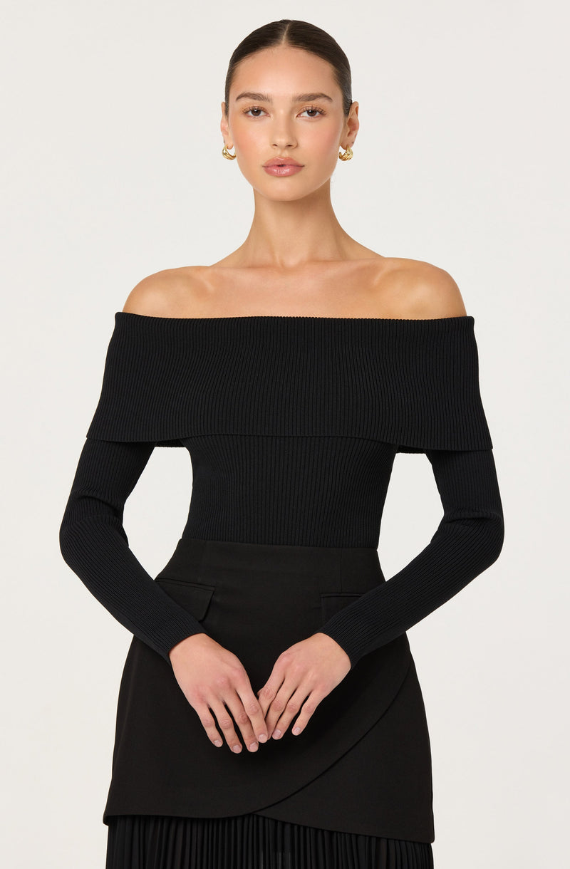 A woman is wearing an off-the-shoulder, long-sleeve black ribbed sweater with a wide neckline that drapes over her shoulders. The sweater fits snugly and has a textured, vertical ribbed pattern. She is also wearing a high-waisted black skirt with subtle darting and layer details, paired with gold earrings and small hoops.