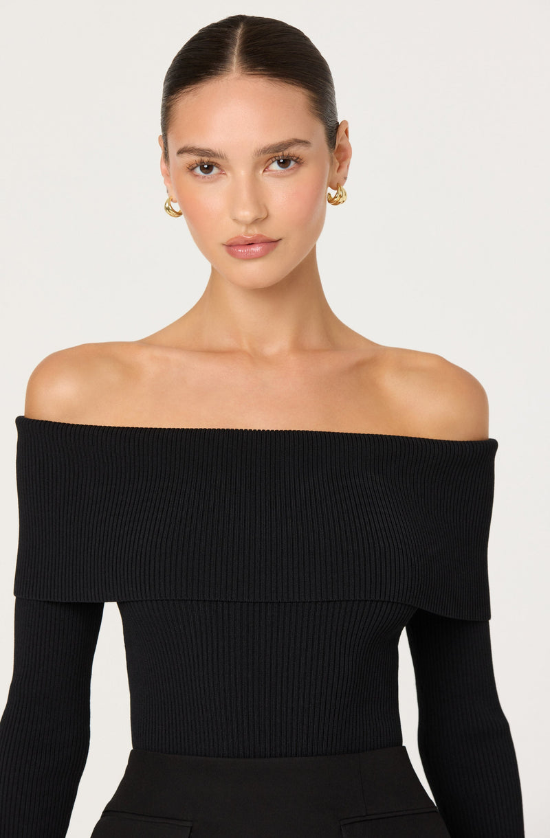 A woman is wearing a black, ribbed, off-the-shoulder top with a wide neckline that drapes over her shoulders. The top has long sleeves and appears fitted. She is accessorized with gold hoop earrings.