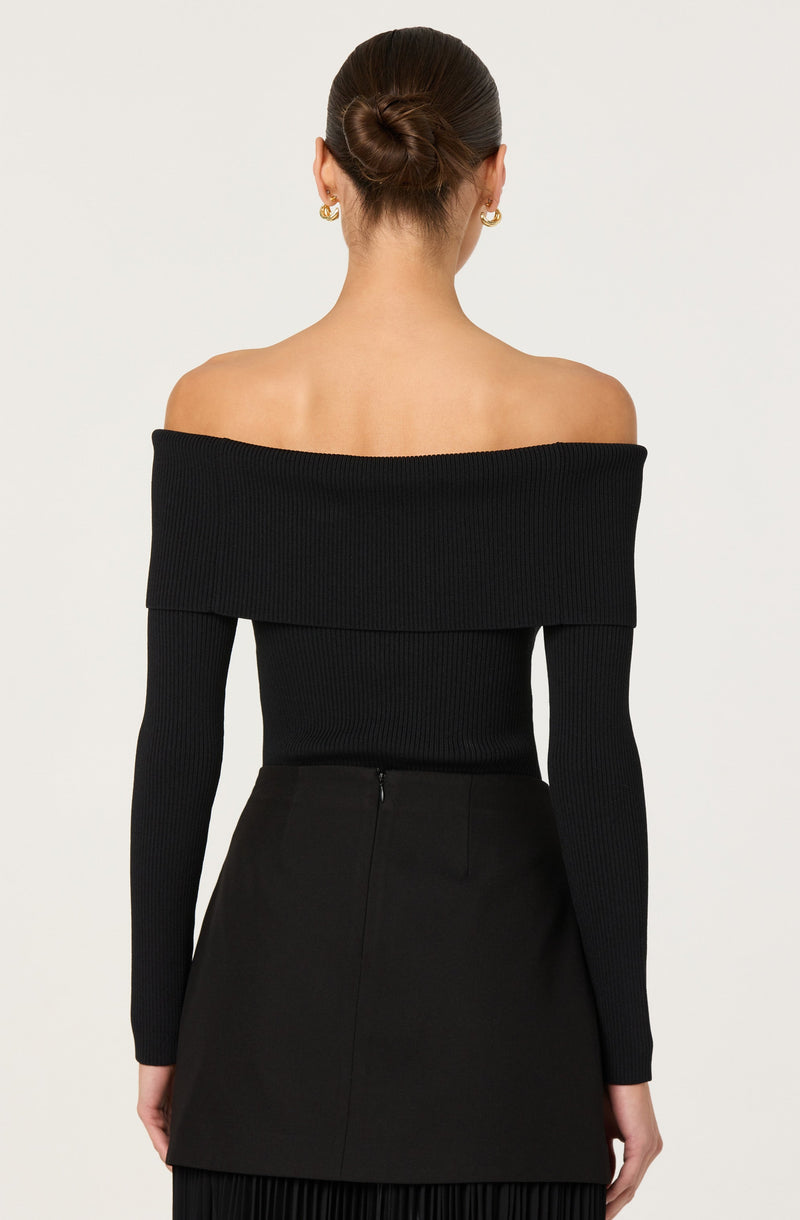 A woman is wearing an off-the-shoulder black ribbed sweater with long sleeves and a wide folded collar that drapes over her shoulders. She has her hair styled in a neat bun and is accessorized with gold hoop earrings.