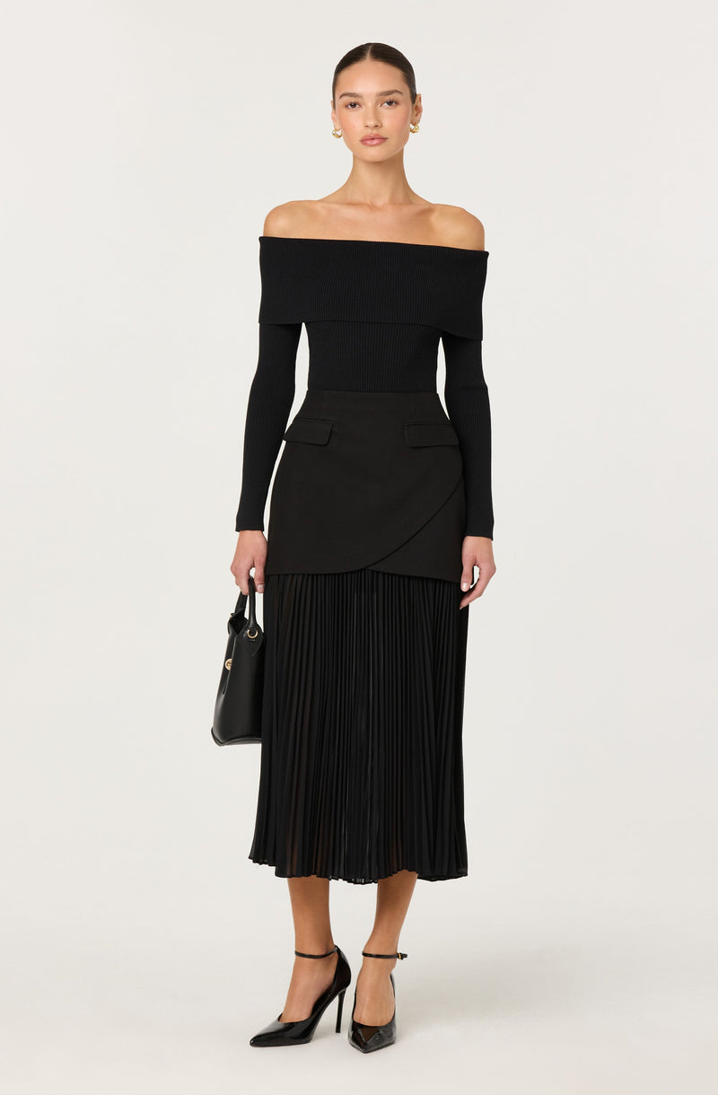 A woman is wearing an off-the-shoulder black top with long sleeves and a wide neckline that exposes her shoulders and collarbone. She pairs it with a black midi skirt that has a wrap design and pleated details at the hem. She is also wearing black high-heeled shoes with ankle straps and is carrying a black handbag.