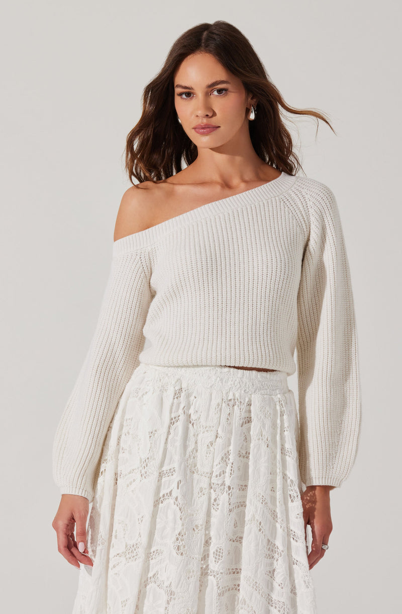 A woman is wearing an off-the-shoulder, cream-colored knit sweater with a ribbed pattern. The sweater has long, slightly puffed sleeves and is cropped at the waist. She pairs it with a high-waisted, white lace skirt that features a delicate floral pattern and a flowing silhouette.
