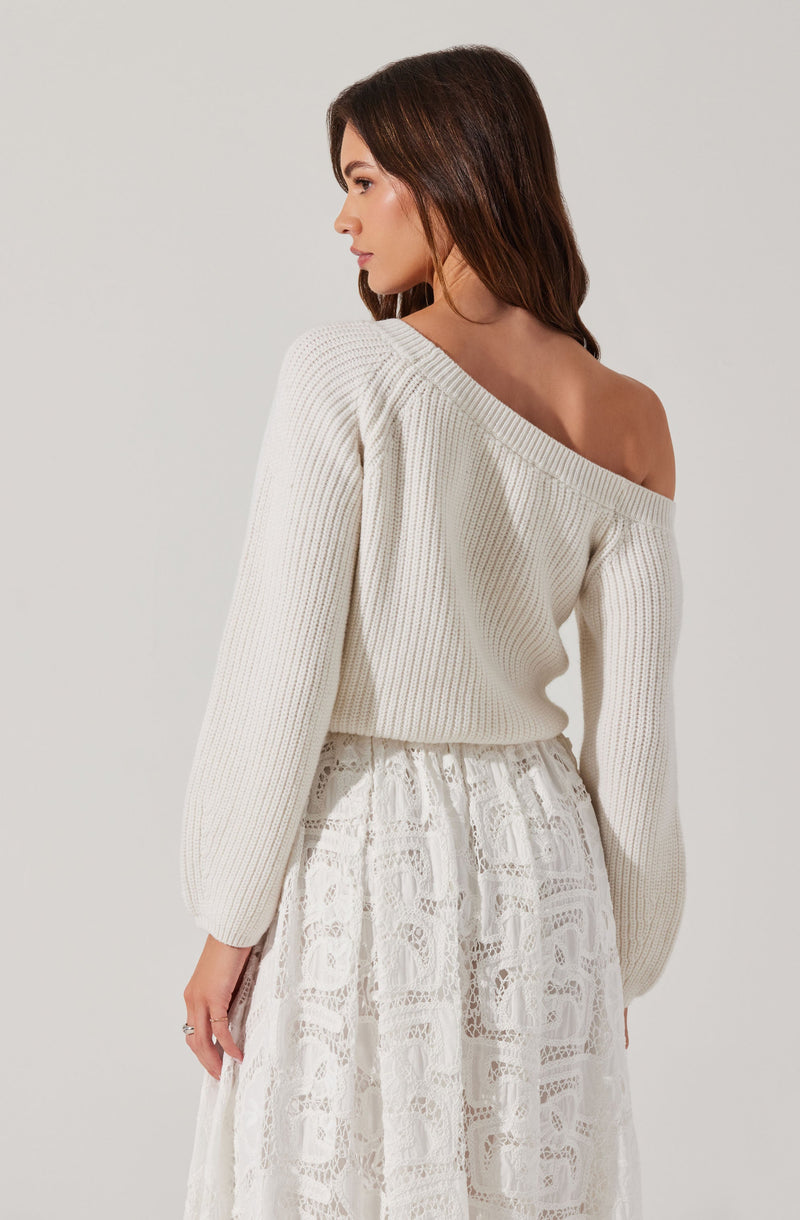 A woman is wearing an off-the-shoulder, cream-colored, oversized knit sweater with long sleeves. She is also dressed in a white, lace, midi-length skirt with intricate patterns.