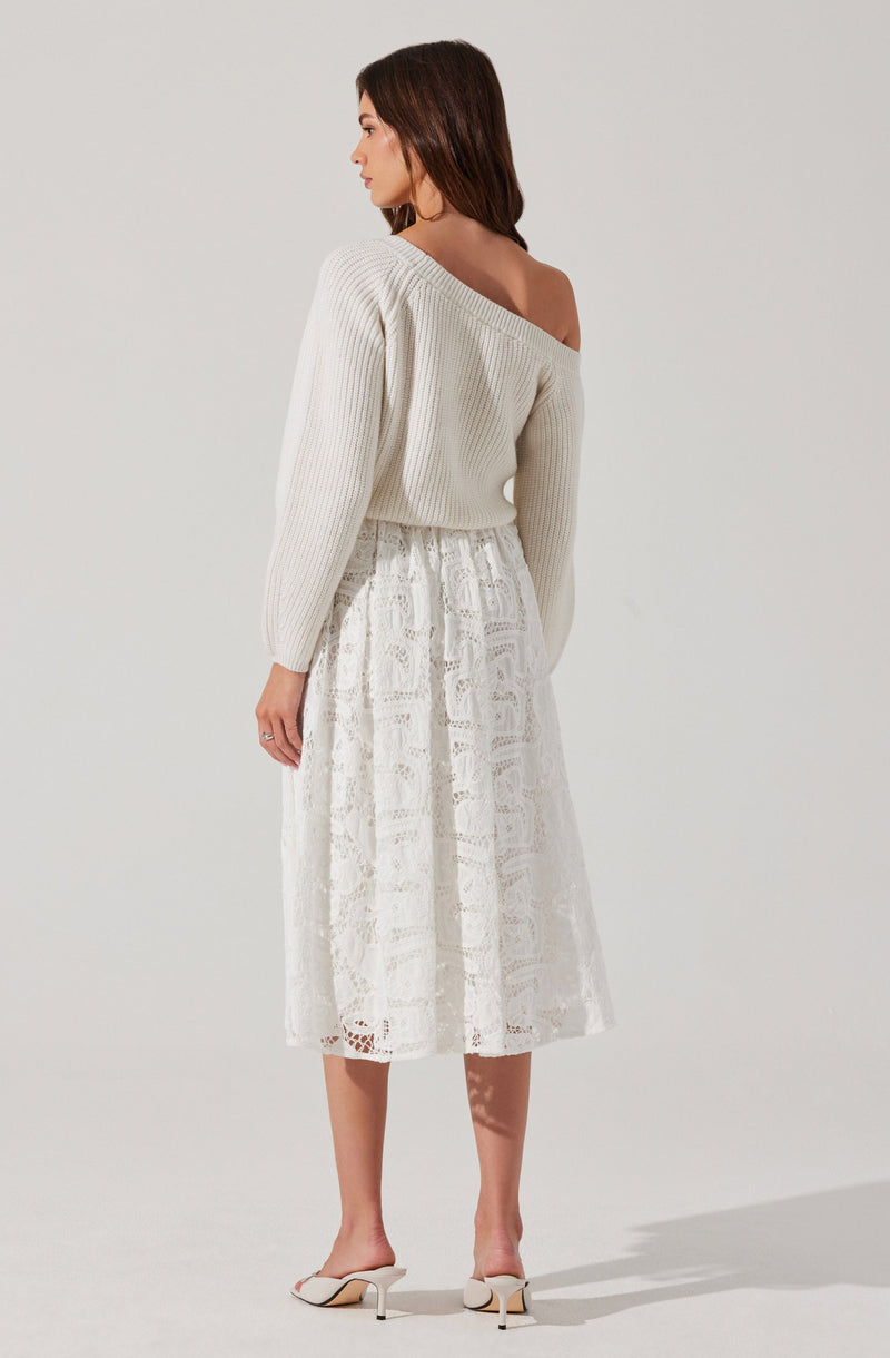 A woman is wearing an off-the-shoulder, long-sleeved, cream-colored, ribbed knit sweater paired with a white, ankle-length lace skirt featuring intricate embroidery and mirror work. She is also wearing white high-heeled shoes with ankle straps.