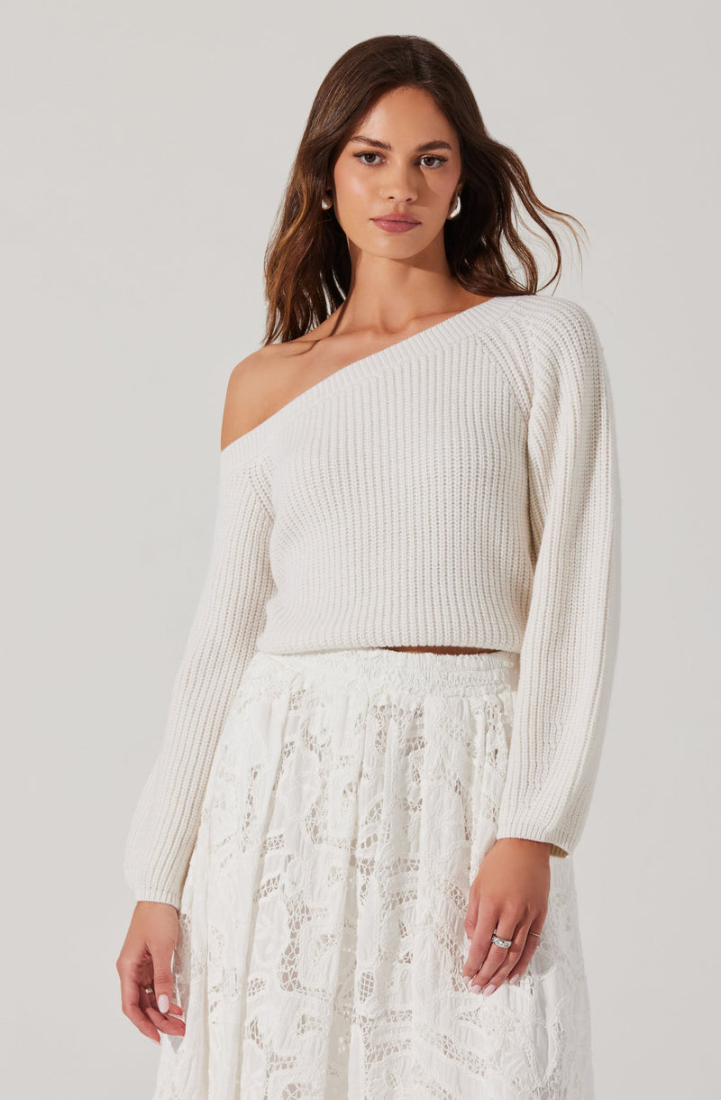 A woman is wearing a cream-colored, off-the-shoulder knitted sweater with long sleeves, paired with a white lace skirt that has intricate floral patterns.
