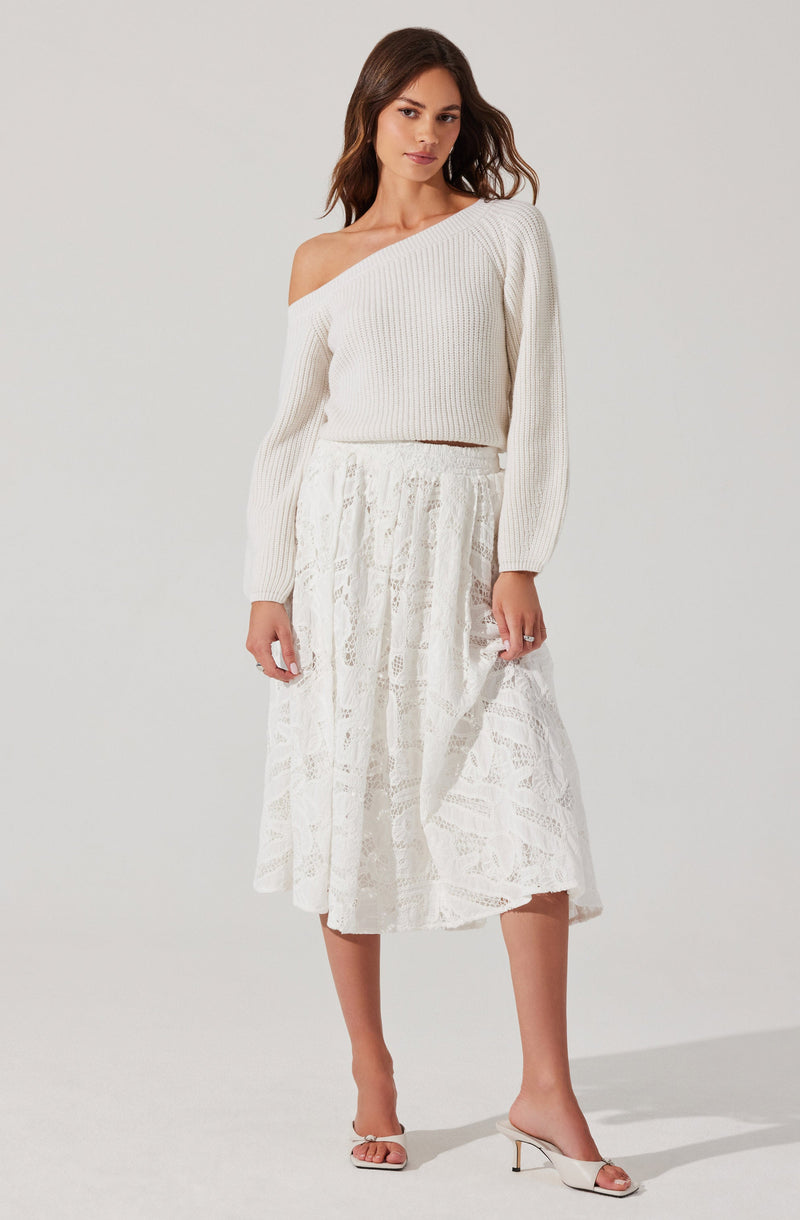 A woman is wearing an off-the-shoulder cream-colored knitted sweater with long sleeves and a loose fit. She pairs it with a white, lace, midi-length skirt that has a soft, flowy silhouette and intricate lace detailing. She completes the outfit with light-colored, open-toe heeled sandals.