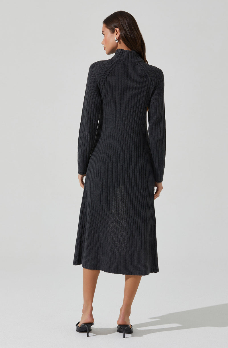 A woman is wearing a long, ribbed knit dress in a dark color, possibly black or charcoal gray. The dress features a high neckline and long sleeves, with a fitted silhouette that extends to mid-calf length. She is also wearing black open-toe heels.