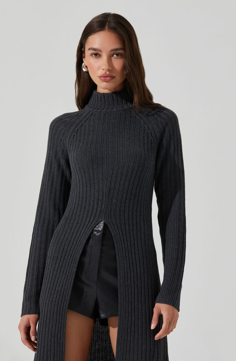 A woman is wearing a long, dark gray, ribbed knit sweater with a high neckline. The sweater features a front slit from the waist down, revealing black shorts underneath. The sweater has a fitted silhouette with long sleeves.