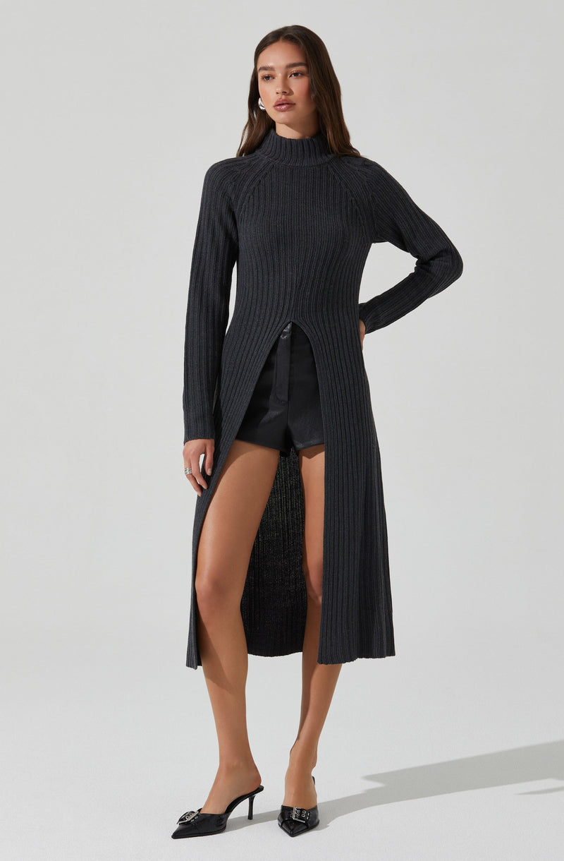 The model is wearing a long, dark gray, ribbed knit sweater with a high neckline. The sweater has a body-hugging fit on the upper part and features a front slit that reveals black shorts underneath. She is also wearing black pointed-toe high heels with decorative embellishments.