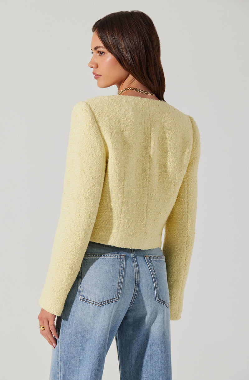A woman is wearing a textured, light yellow, long-sleeved blazer with a cropped length. She pairs it with high-waisted, light blue denim jeans. The outfit is simple yet stylish, with the blazer adding a touch of softness to the casual denim look.