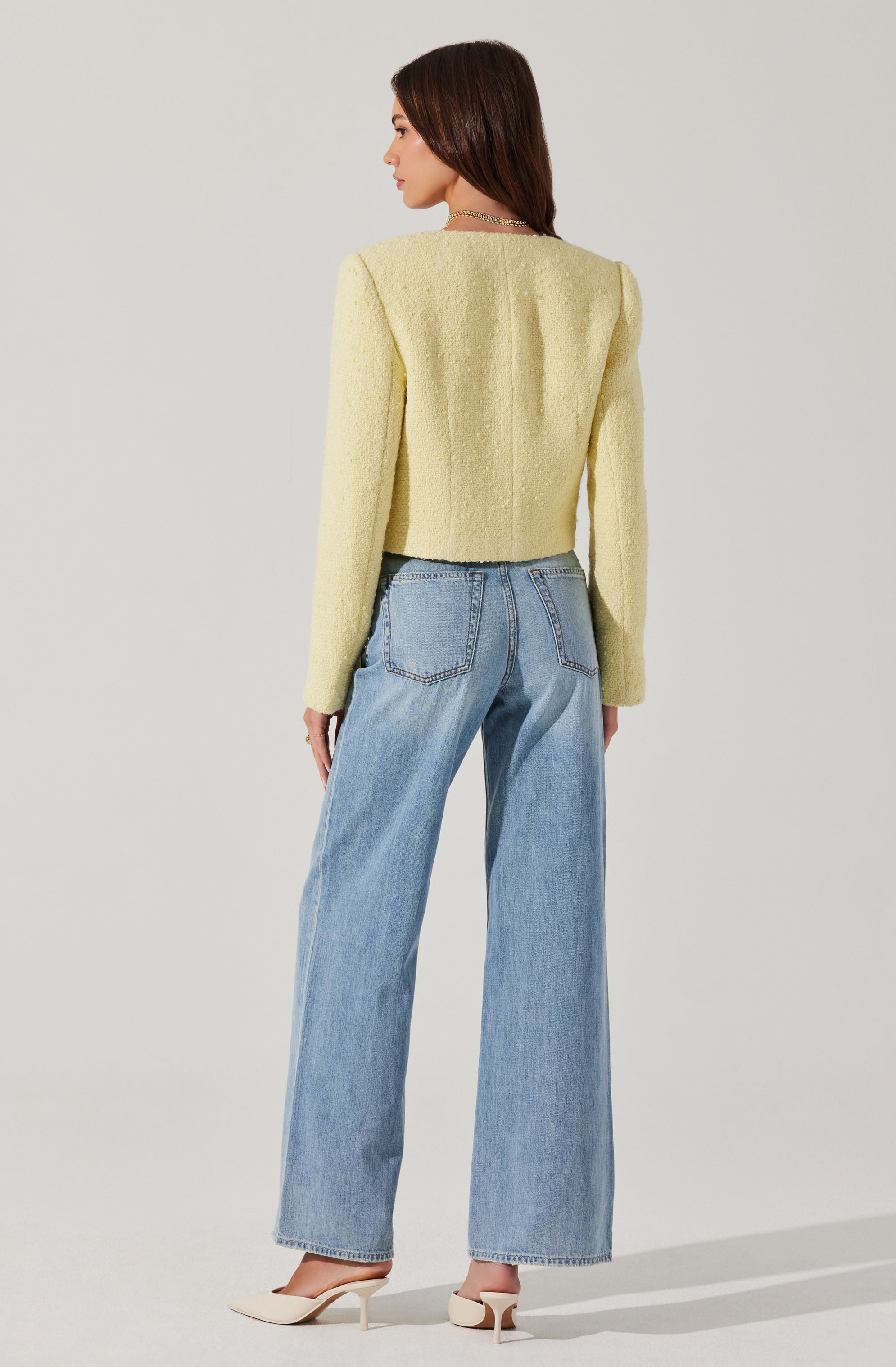 A woman is wearing a light yellow textured cropped blazer with long sleeves and structured shoulders, paired with high-waisted, wide-leg light blue jeans. She is also wearing pointed cream-colored high heels.