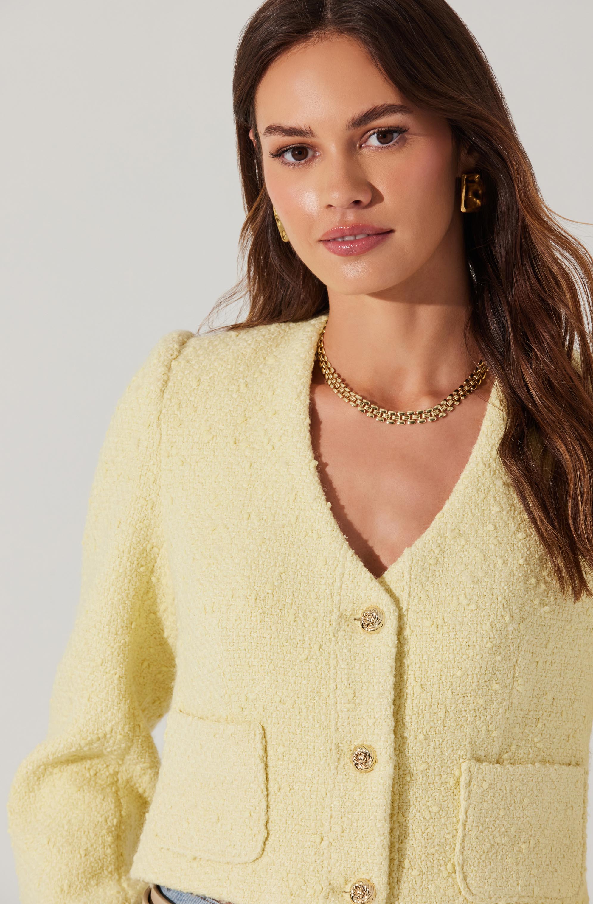 A woman is wearing a pale yellow, textured, long-sleeve cardigan with large gold buttons down the front. She accessorizes with a chunky gold chain necklace and matching gold earrings.
