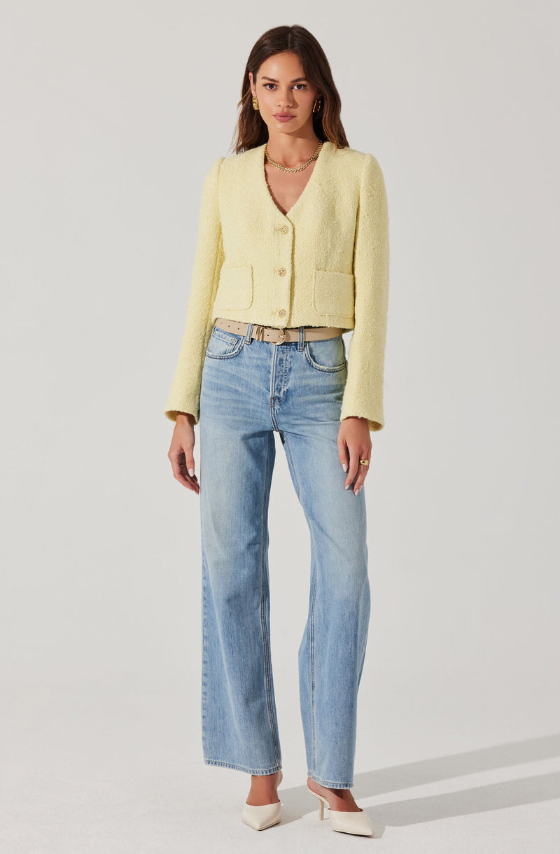 A woman is wearing a light yellow textured, cropped cardigan with three large buttons down the front and two patch pockets at the waist. She has paired it with light blue, high-waisted, straight-leg jeans and white pointed-toe heels. She accessorizes with gold hoop earrings, a gold chain necklace, and a beige belt with a gold buckle.