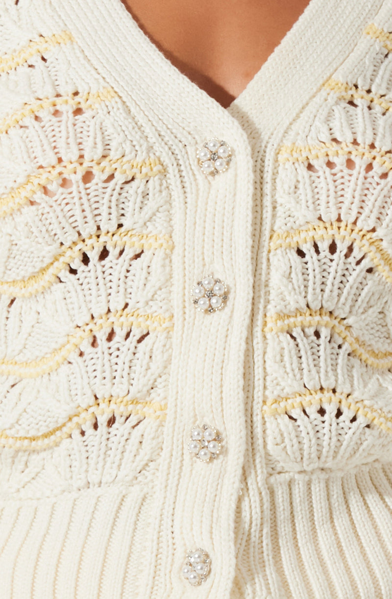 An off-white, knitted cardigan featuring decorative pearl and rhinestone buttons. The cardigan has a textured, open-weave pattern with horizontal stripes of different knit styles. The buttons are round with a floral design made of small pearls and rhinestones. The neckline is a V-shape.