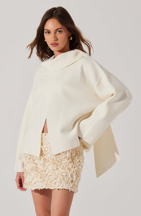 A woman is wearing an oversized, cream-colored sweater with a wide collar that drapes around her shoulders. The sweater has a relaxed fit with dropped shoulders and long sleeves. She pairs it with a cream-colored, textured mini skirt featuring a three-dimensional floral or ruffled pattern.
