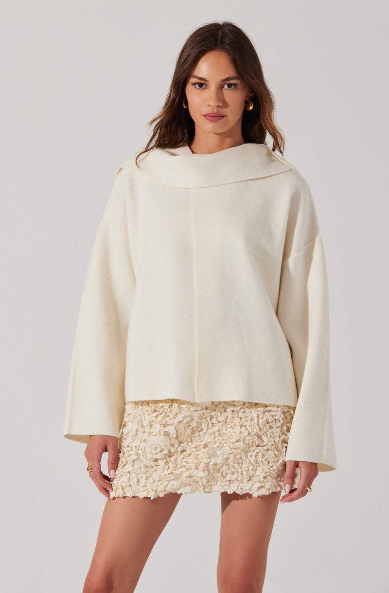 A woman is wearing an oversized, off-white top with a wide, rounded collar and long sleeves. She pairs it with a short, textured cream-colored skirt that has a floral, ruffled pattern.