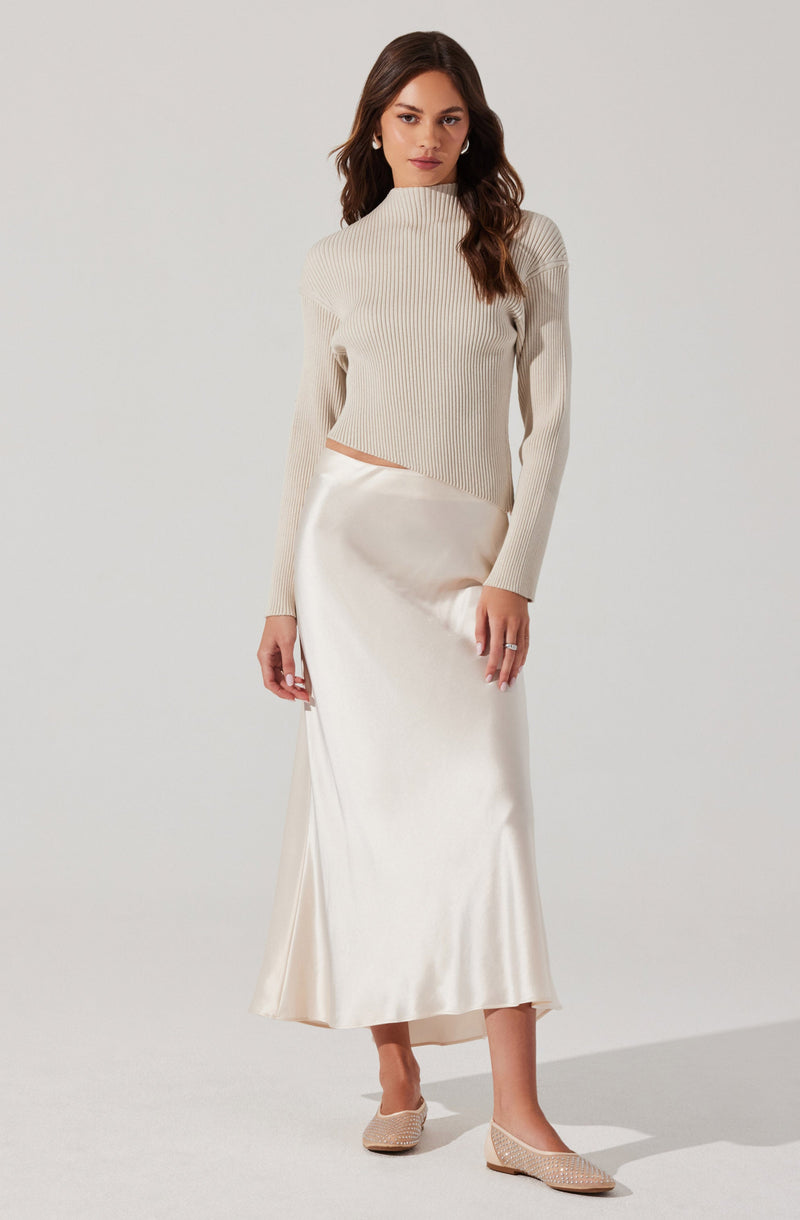 A woman is wearing a cream-colored, ribbed turtleneck sweater with long sleeves, paired with a silky, satin-like cream-colored maxi skirt that reaches her ankles. She completes the outfit with pointed-toe, woven beige flats.