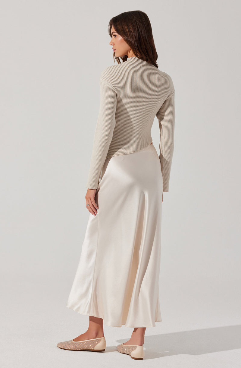 A woman is wearing a light beige, fitted, ribbed long-sleeve sweater with a high neckline. She pairs it with a long, flowing, satin-like cream-colored skirt that reaches her ankles and features a subtle slit on the side. She completes the outfit with beige, pointed-toe ballet flats that have a textured woven pattern.
