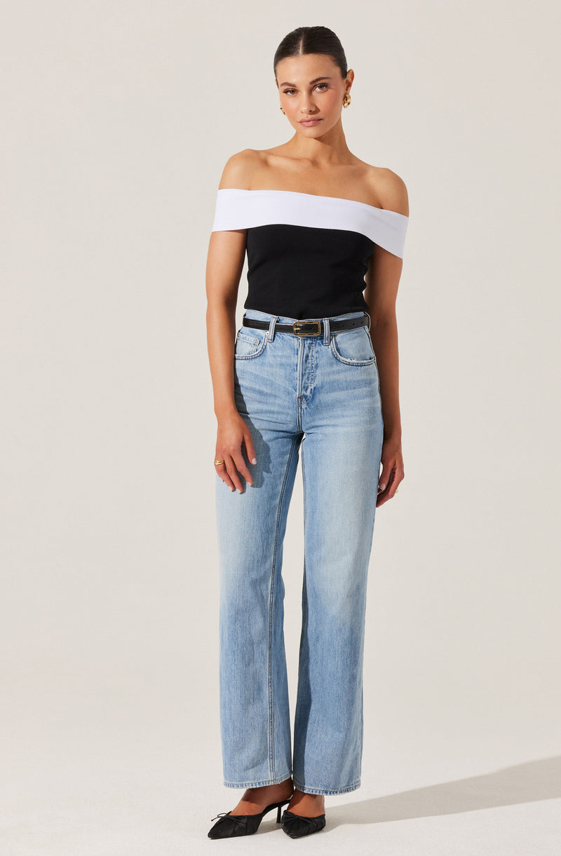 A woman is wearing an off-the-shoulder top with a wide white band across the shoulders and a black body. She has paired it with high-waisted, light-wash denim jeans that have a relaxed, straight-leg fit. The jeans are secured with a black belt with a gold buckle. She is also wearing black pointed-toe heels.