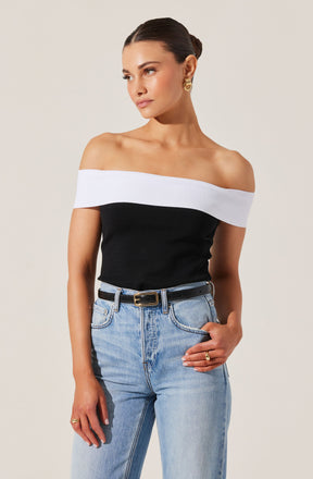 A woman is wearing an off-the-shoulder top with a white band across the shoulders and upper arms, and a black body. She pairs it with high-waisted, light-wash jeans that have a relaxed fit, and a black belt with a gold buckle. She accessorizes with gold hoop earrings and a gold ring on her right hand.