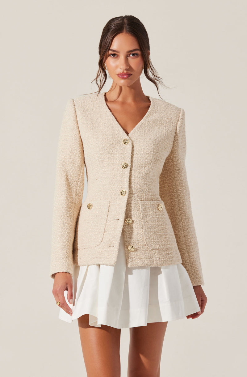 A woman is wearing an elegant cream-colored textured blazer with gold buttons down the front and on the pockets. The blazer has a tailored fit with a V-neckline. She pairs it with a white pleated skirt.