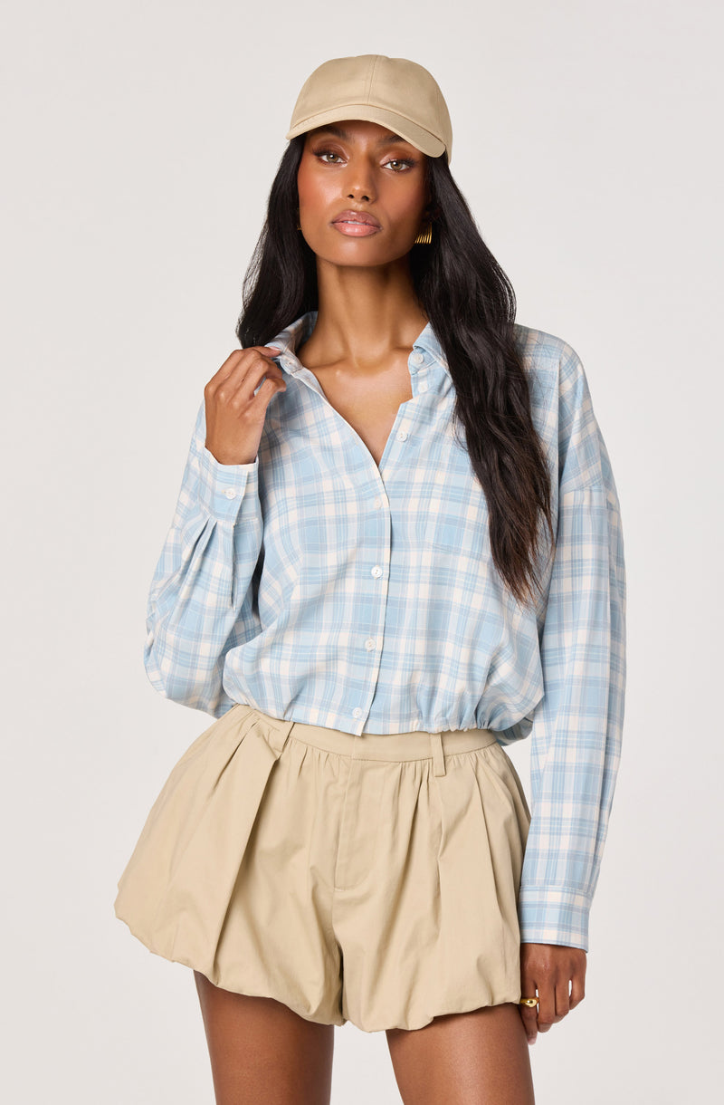 Woman in a light blue plaid shirt and beige pleated shorts, with a beige cap.