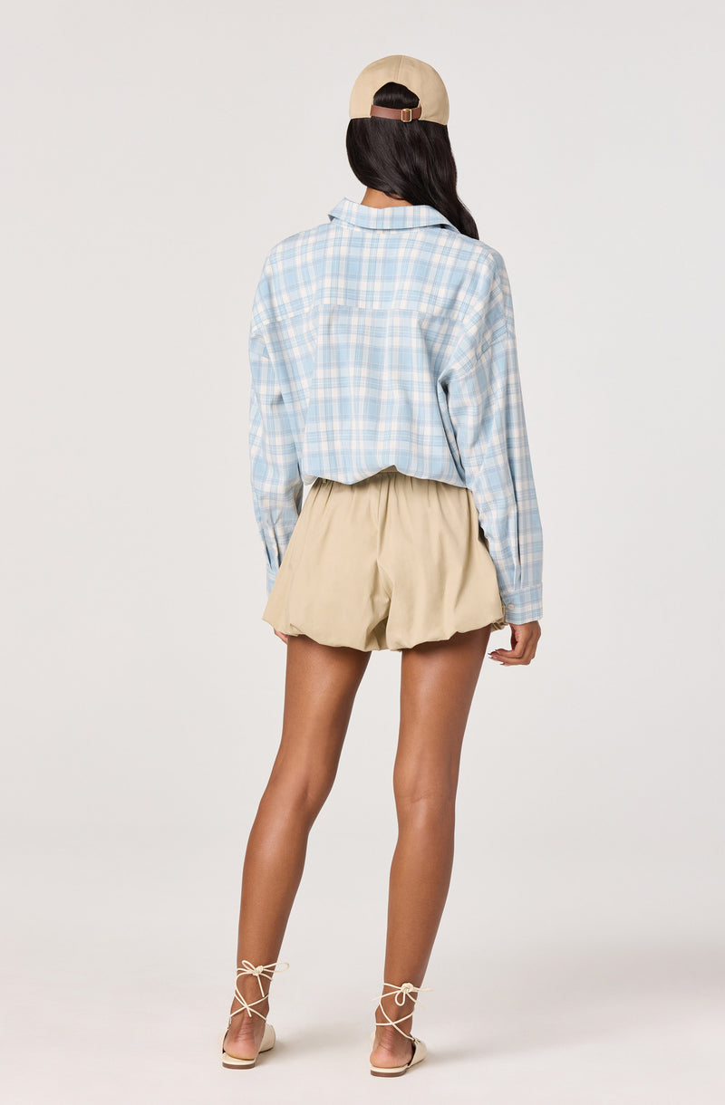 Woman wearing a light blue plaid shirt, beige shorts, and beige sandals with ankle ties, back view.