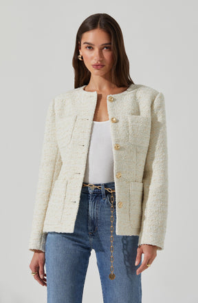 A woman is wearing a cream-colored, textured, cropped blazer with large, gold buttons down the front. The blazer has structured shoulders and patch pockets on the chest. She pairs it with a white top and high-waisted blue jeans, accessorized with a gold chain belt and statement earrings.