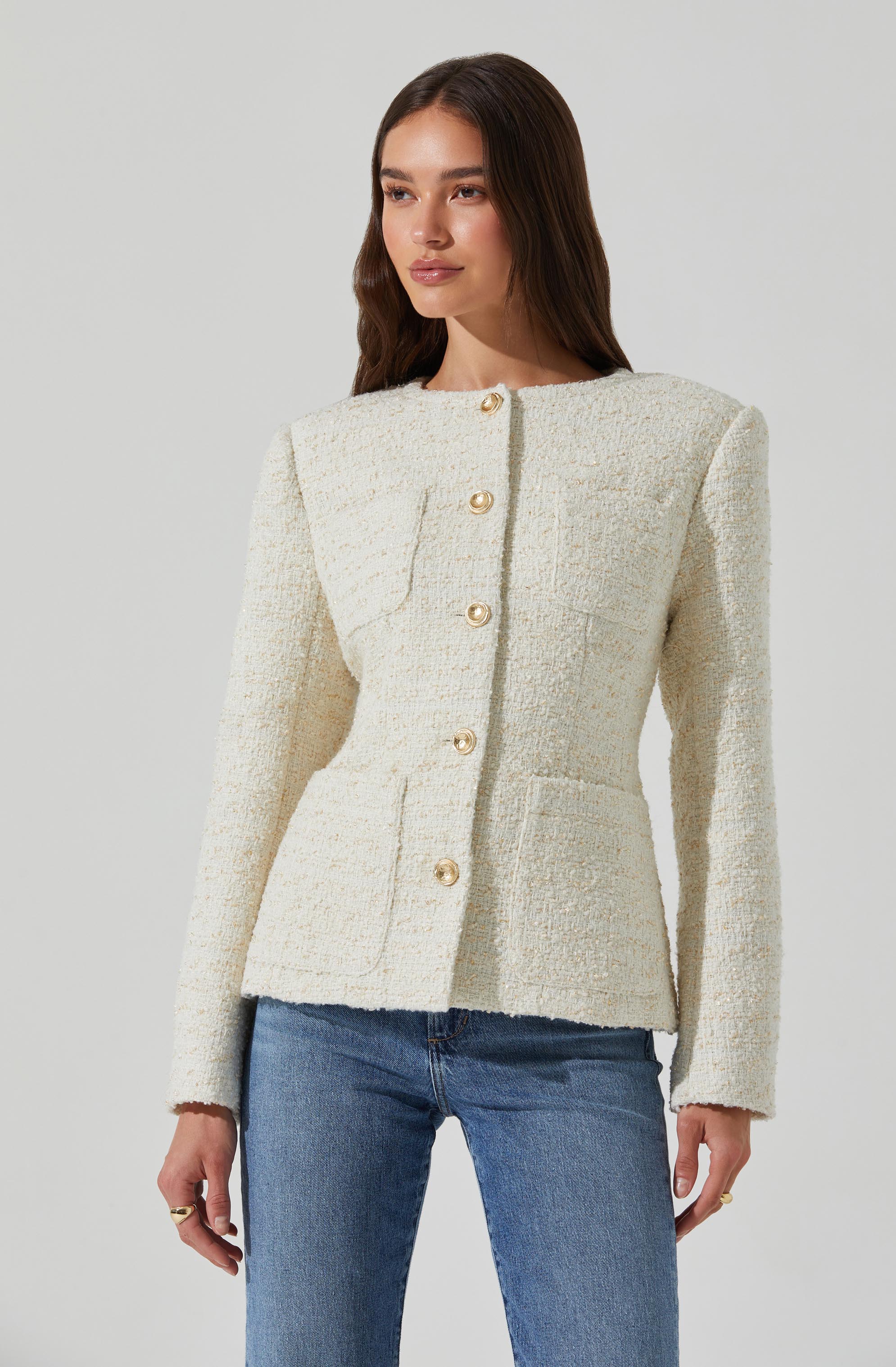 A woman is wearing an ivory textured blazer with a rounded neckline and shoulder pads. The blazer features large, shiny gold buttons down the front and two front pockets. She is also wearing blue jeans.