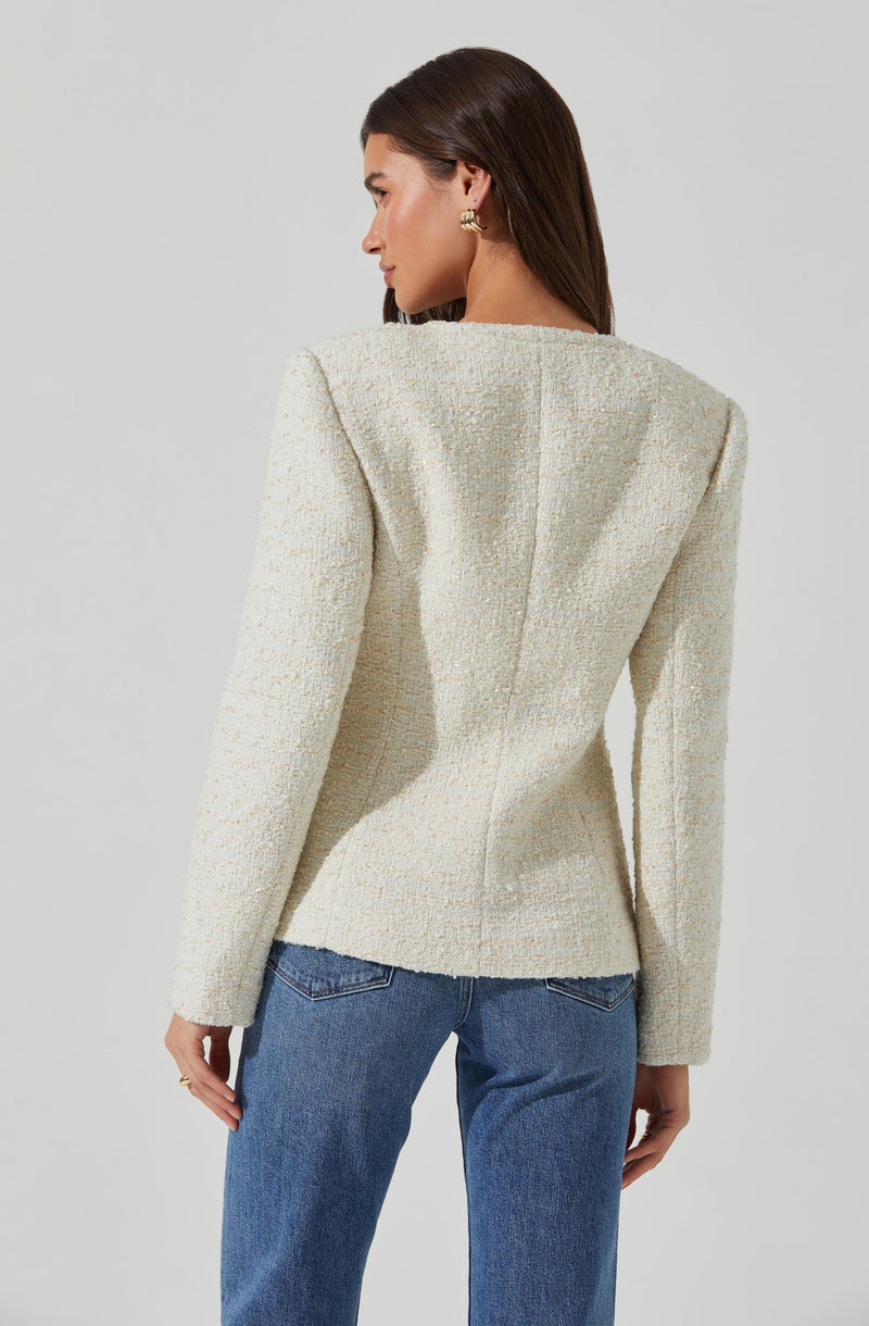 A woman is wearing a fitted, cream-colored textured blazer with structured shoulders and a back seam detail. She pairs it with blue jeans. She has shoulder-length dark brown hair and is accessorized with gold earrings.