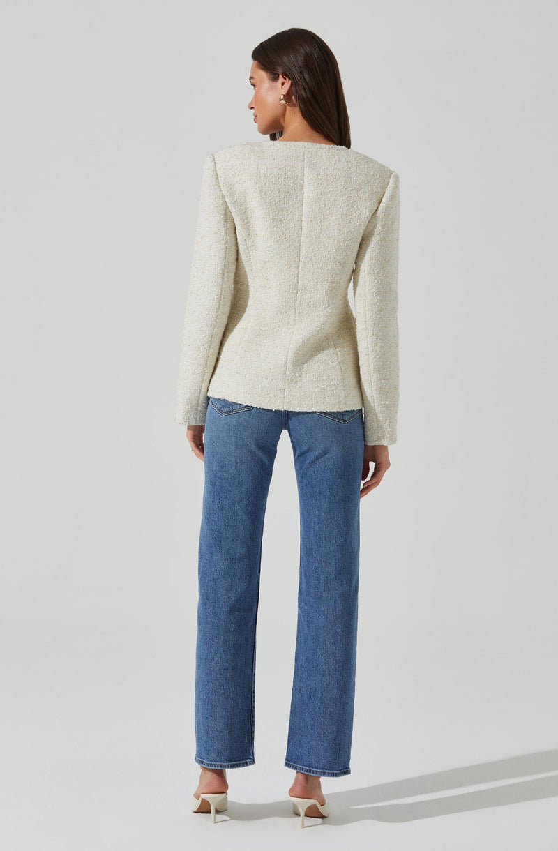 A woman is wearing a cream-colored textured blazer with a structured, slightly padded shoulder design. She pairs it with straight-leg blue jeans and cream high-heeled shoes.