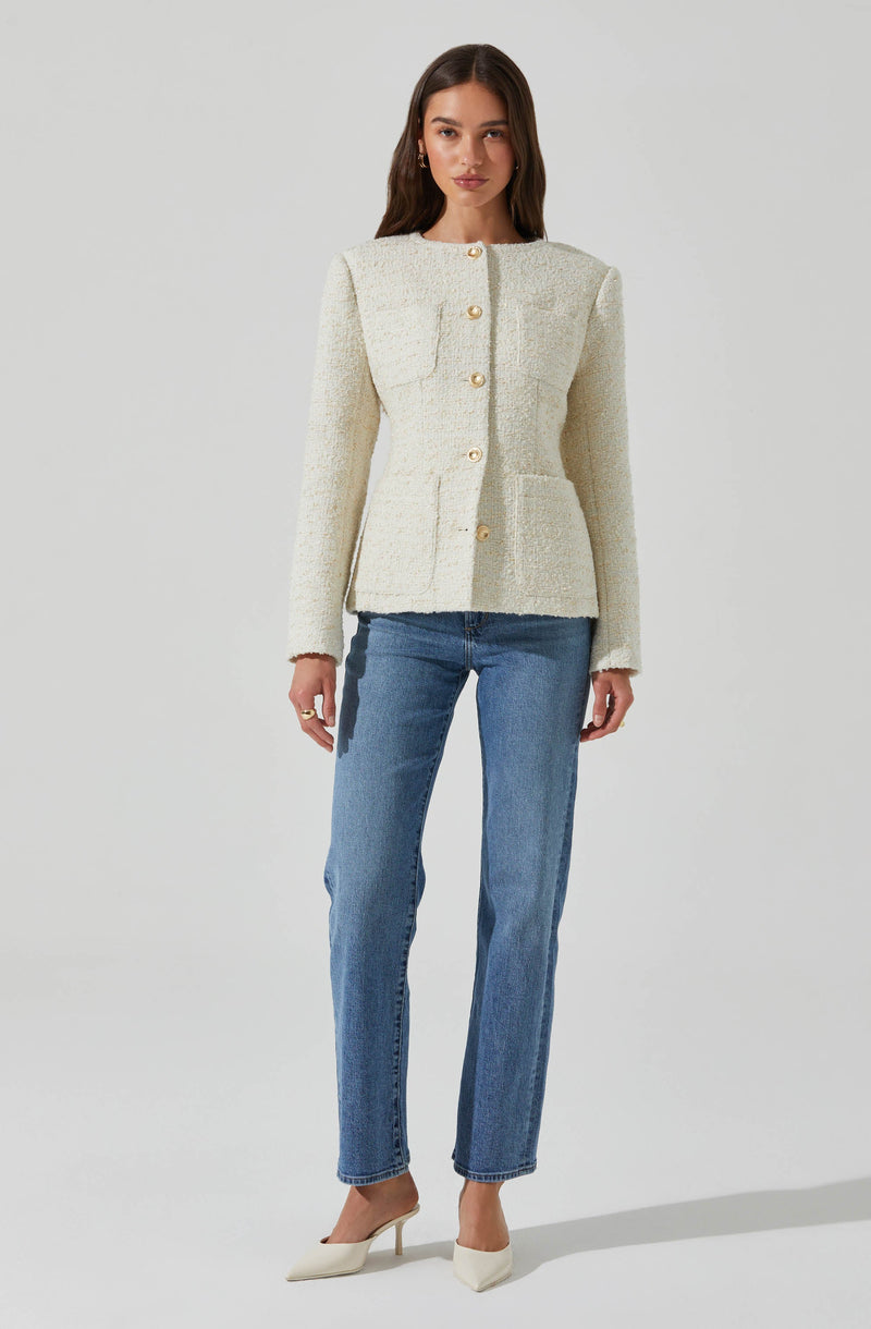A woman is wearing a cream-colored textured blazer with a round neckline and large gold buttons down the front. The blazer has structured shoulders and two front patch pockets. She pairs it with high-waisted, light-wash straight-leg jeans and white pointed-toe heels.