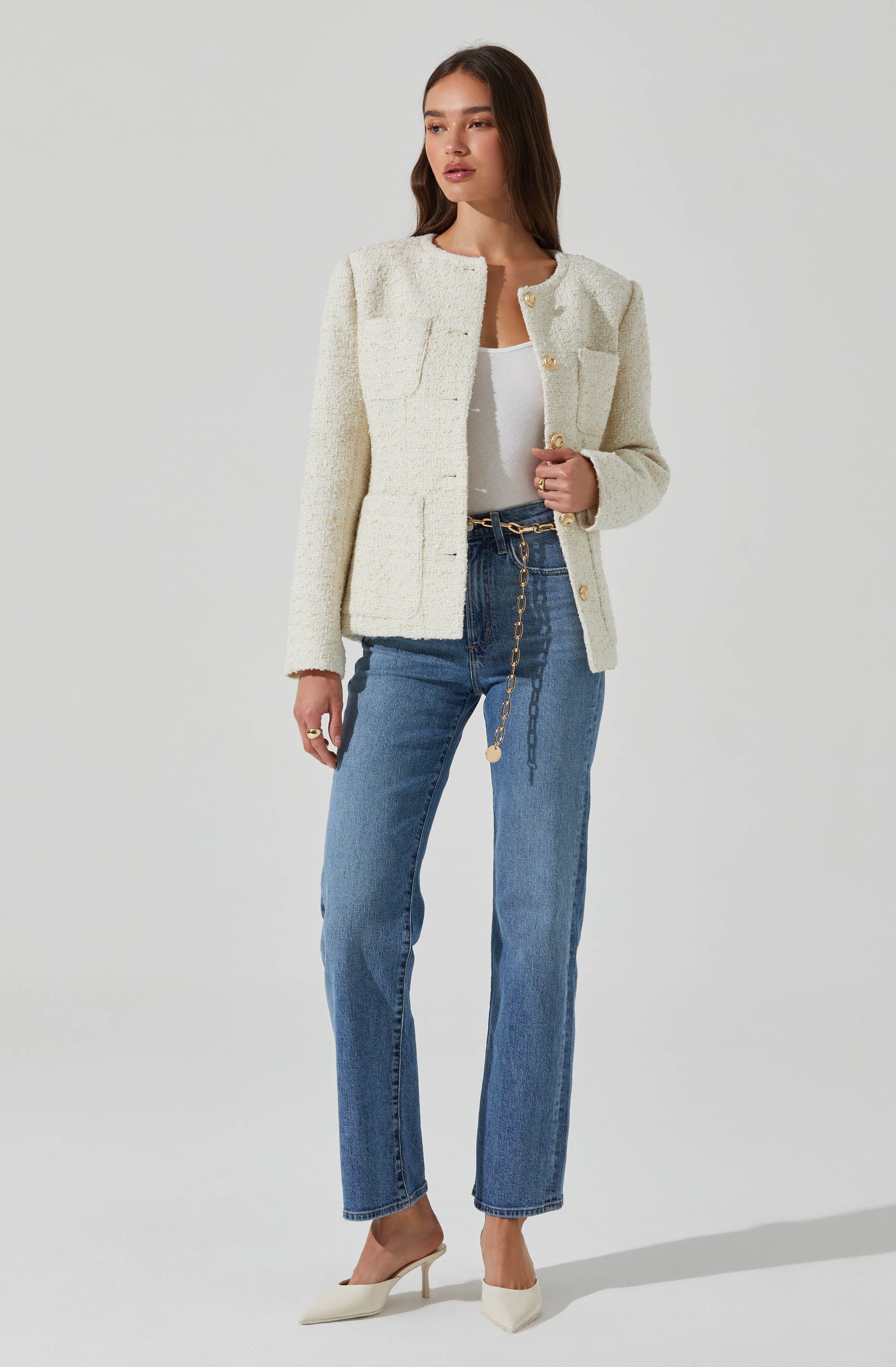 A woman is wearing a cream-colored, textured, long-sleeve, collarless jacket with gold buttons down the front and on the pockets. She has on a plain white top underneath. She is also wearing high-waisted, straight-leg blue jeans with a chain belt hanging from the waistband. Her shoes are pointed-toe, cream-colored high heels.