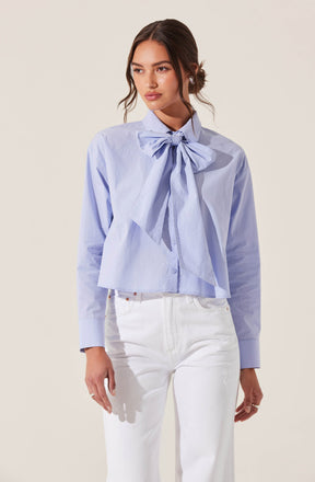 A woman is wearing a light blue button-up shirt with a large, decorative bow tied at the collar. The shirt has long sleeves with buttoned cuffs and a slightly cropped, loose fit. She pairs the shirt with high-waisted white jeans.