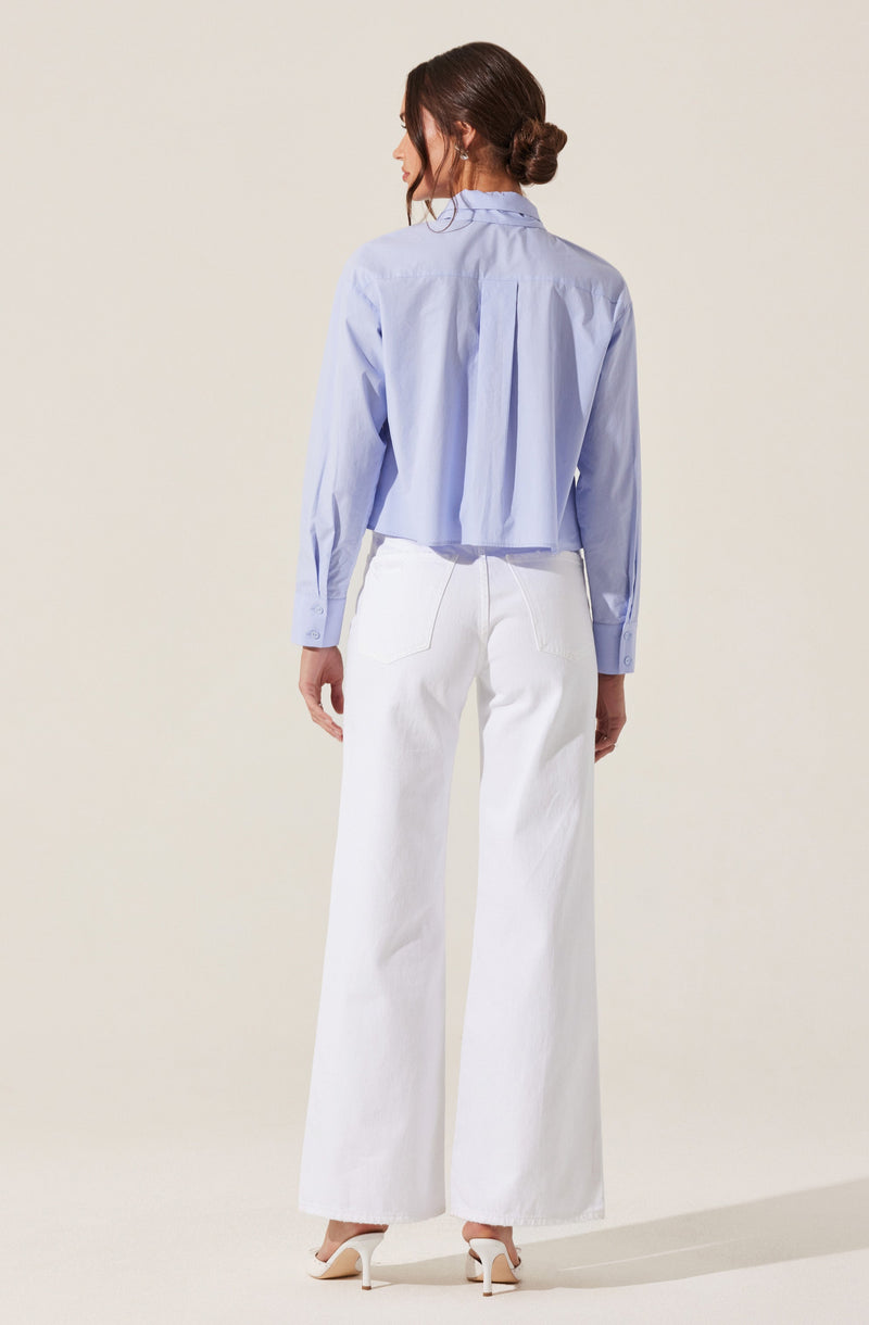 A woman is wearing a light blue long-sleeved button-up shirt with a collar and pleats at the back. She is also wearing high-waisted, wide-leg white pants and beige high-heeled shoes. Her hair is styled in a loose bun.