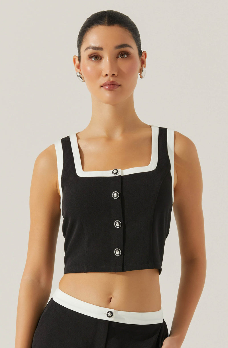 A woman is wearing a sleeveless, black cropped top with wide straps and a square neckline outlined by a white border. The top features four large, round buttons down the front. She is also wearing black high-waisted pants accented with a white waistband, which has a decorative button in the center. She has her hair styled in a sleek, pulled-back look and is accessorized with hoop earrings. The background is plain and light-colored.