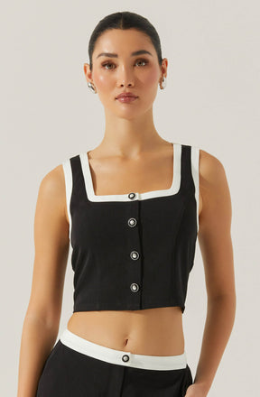 A woman is wearing a sleeveless, black cropped top with wide straps and a square neckline outlined by a white border. The top features four large, round buttons down the front. She is also wearing black high-waisted pants accented with a white waistband, which has a decorative button in the center. She has her hair styled in a sleek, pulled-back look and is accessorized with hoop earrings. The background is plain and light-colored.